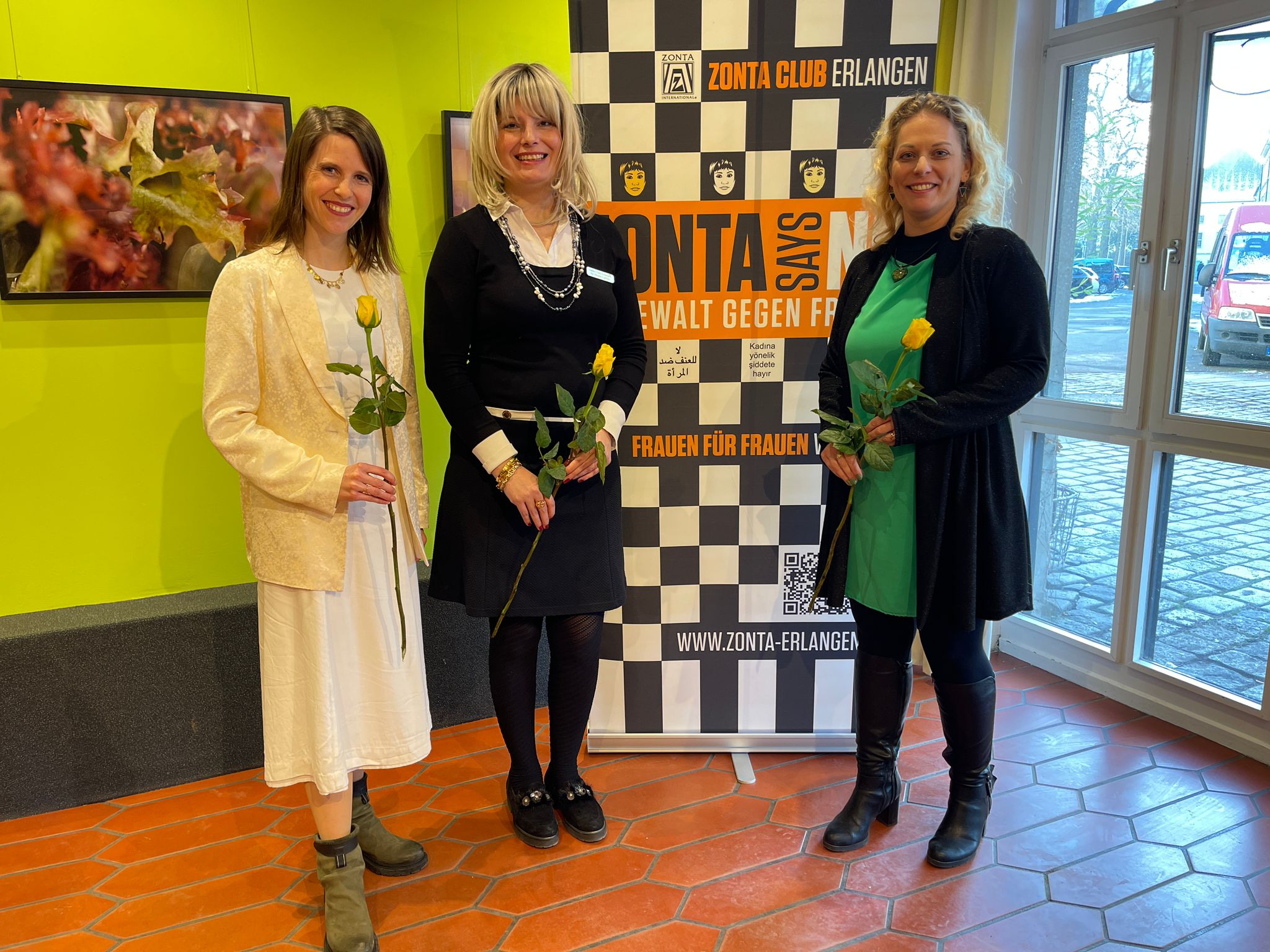 Eva Gengler, Dr. Magda Luthay und Cindy Uslu stehen mit je einer gelben Rose in der Hand vor dem Aufsteller des ZONTA Club Erlangen